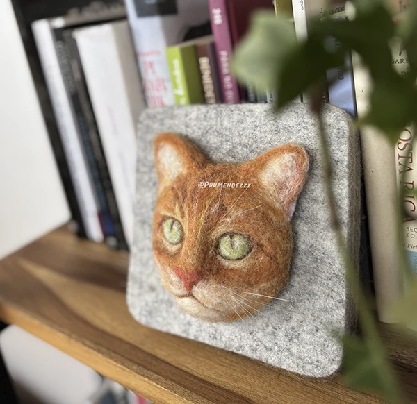 Felt picture of an orange cat's face resting on a shelf
